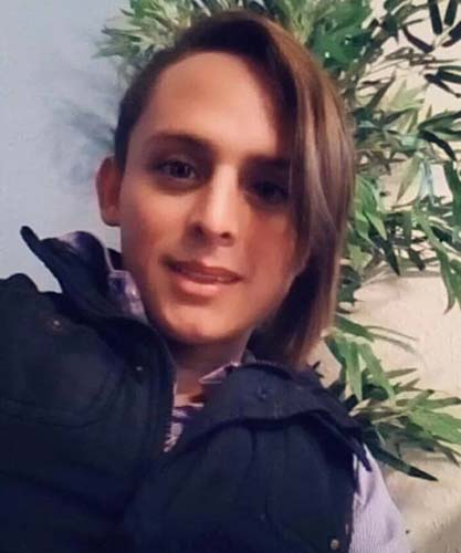 A young trans woman looks into the camera. She is wearing a dark blue coat, has olive skin, and light brown hair, shaved on the right side and long on the left. Behind her is a tall, green house plant.
