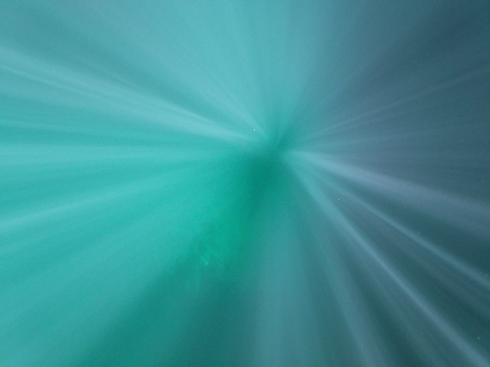 Vivid blues and greens are cut through by blurry rays of light shimmering out from the center, giving the impression of looking down into a watery expanse.